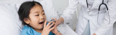 asian child holding inhaler and opening mouth near doctor in white coat in hospital ward, banner