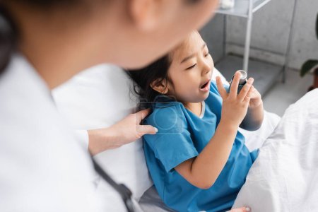 Foto de Pediatra borrosa tocando hombro de niño asiático usando inhalador en sala de hospital - Imagen libre de derechos