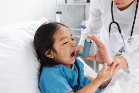 Asiatin öffnet Mund in der Nähe von Arzt mit Inhalator in Kinderklinik