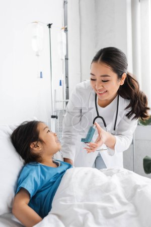 smiling doctor holding inhaler near positive girl on bed in hospital ward