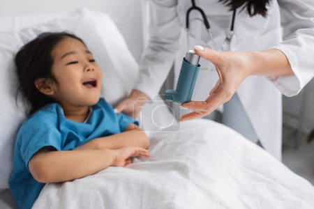 verschwommener Kinderarzt hält Inhalator in der Nähe von asiatischem Kind, das Mund auf Krankenhausbett öffnet