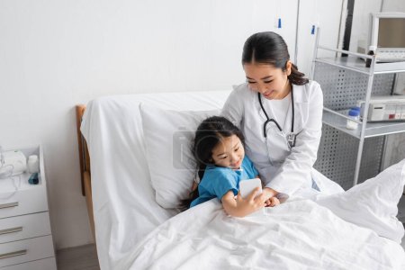 Téléchargez les photos : Enfant insouciant prendre selfie sur smartphone avec sourire asiatique médecin dans la salle d'hôpital - en image libre de droit