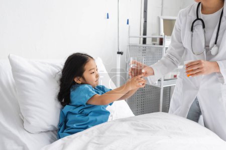 Arzt im weißen Mantel hält Tablettenbehälter und gibt asiatischem Mädchen ein Glas Wasser auf dem Krankenhausbett