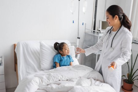 Lächelnder Kinderarzt mit Tablettenbehälter und Wasserglas in der Nähe unbeschwerter Asiatin auf dem Bett in der Klinik