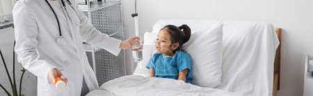 Arzt in weißem Mantel mit Medikamenten und einem Glas Wasser in der Nähe eines asiatischen Mädchens, das auf dem Krankenhausbett lächelt, Transparent