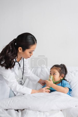 Téléchargez les photos : Jeune médecin aider asiatique fille respiration dans oxygène masque sur lit dans hôpital salle - en image libre de droit