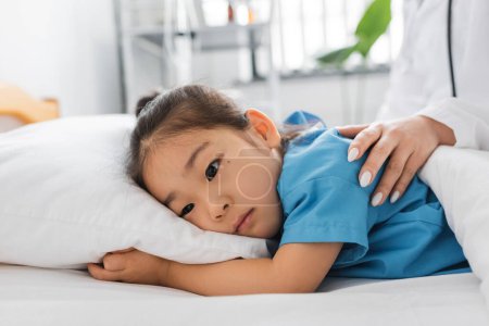 Téléchargez les photos : Médecin calmant bouleversé asiatique fille couché sur lit dans hôpital salle - en image libre de droit
