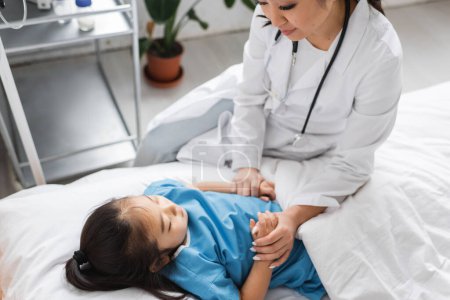 asiatischer Arzt in weißem Mantel hält Hände einer kleinen Patientin, die in Kinderklinik auf dem Bett liegt