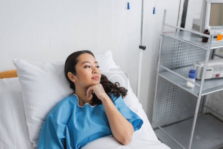 mujer asiática soñadora sosteniendo la mano cerca de la barbilla y mirando hacia otro lado en la cama en el hospital