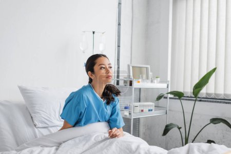serious and thoughtful asian woman in hospital gown sitting on bed in clinic and looking away