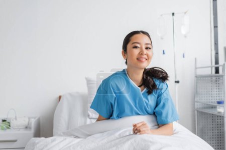 Foto de Alegre asiático mujer sentado en cama en hospital sala y sonriendo a cámara - Imagen libre de derechos