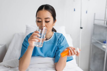junge Asiatin trinkt Wasser, während sie Tablettenbehälter auf dem Bett in der Klinik hält