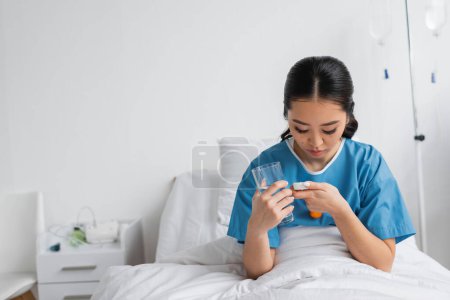 junge Asiatin schaut in Tablettenbehälter, während sie ein Glas Wasser auf dem Krankenhausbett hält