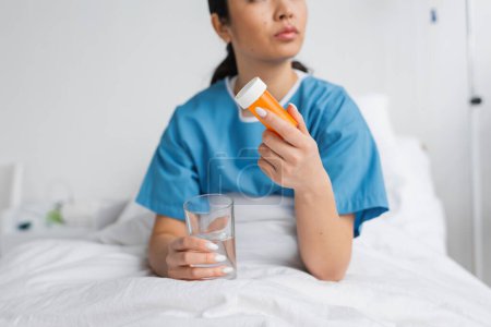 partial view of woman in hospital gown holding water and pills container on bed in clinic