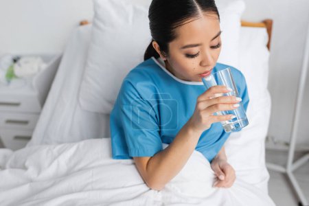 brunette asian woman sitting on bed in hospital ward and drinking fresh water