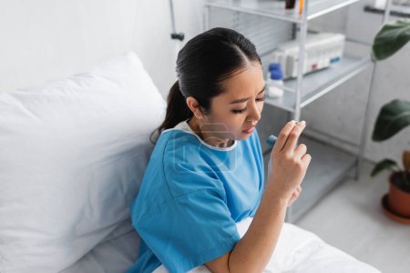 high angle view of sick asian woman holding inhaler while sitting on bed in hospital ward