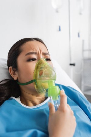 Téléchargez les photos : Inquiet asiatique femme respiration dans oxygène masque tandis que couché sur lit dans clinique - en image libre de droit