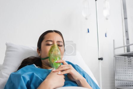 sick asian woman breathing in oxygen mask while lying with closed eyes on bed in clinic