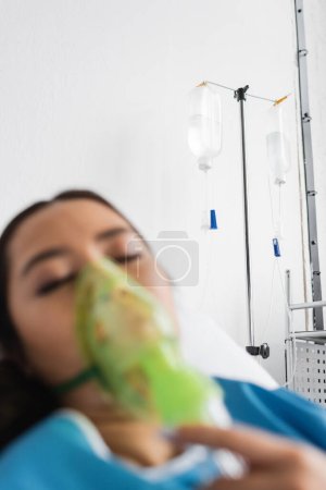 verschwommene Asiatin mit geschlossenen Augen atmet in Sauerstoffmaske auf Krankenhausstation