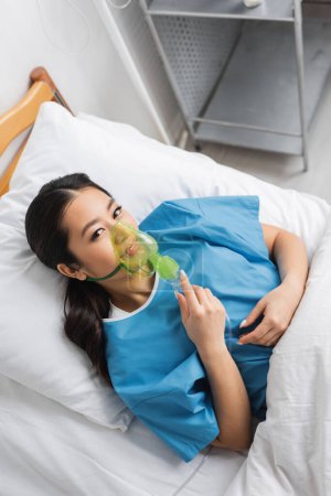 top view of positive asian woman breathing in oxygen mask and looking at camera on bed in clinic