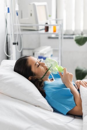 Foto de Vista lateral de la mujer asiática enferma sosteniendo máscara de oxígeno mientras está acostado en la cama en la sala de hospital - Imagen libre de derechos