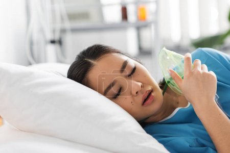 diseased asian woman with closed eyes lying on hospital bed with oxygen mask