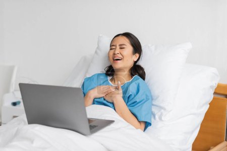 Téléchargez les photos : Excité asiatique femme toucher poitrine et rire avec les yeux fermés tout en regardant film comédie sur ordinateur portable dans clinique - en image libre de droit