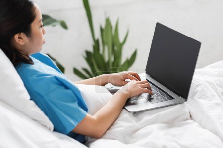 Foto de Joven asiático mujer sentado en hospital cama y mecanografía en portátil con blanco pantalla - Imagen libre de derechos