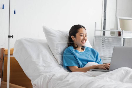 alegre asiático mujer mecanografía en portátil mientras permaneciendo en hospital cama