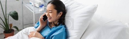 freudige asiatische Frau telefoniert mit Handy und lächelt mit geschlossenen Augen auf dem Bett in der Klinik, Banner