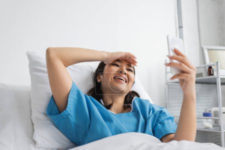 Foto de Excitada mujer asiática tocando la frente y mirando el teléfono inteligente en la cama de hospital - Imagen libre de derechos