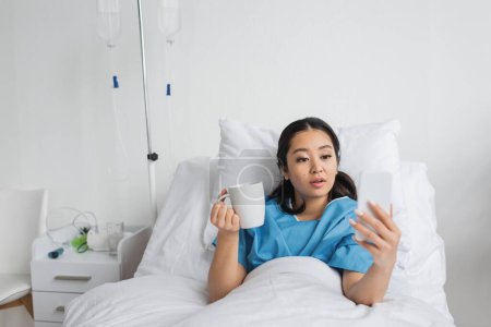 Foto de Mujer asiática sorprendida sosteniendo taza de té y mirando el teléfono móvil en la cama en la clínica - Imagen libre de derechos