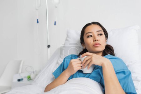 Foto de Pensativo asiático mujer holding té taza y mirando lejos en hospital cama - Imagen libre de derechos