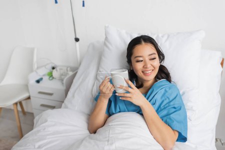 pleased asian woman lying on hospital bed with cup of tea and looking away