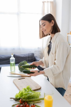 Lächelnde Frau hält Gurken neben Gemüse und Schneidebrett in der Küche 
