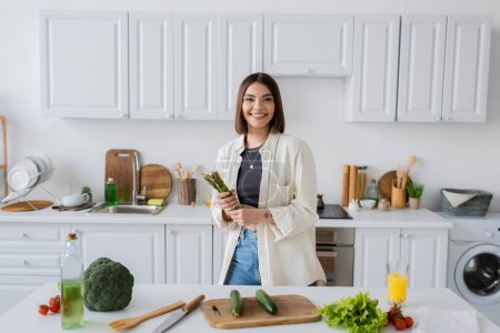 Cheerful woman holding asparagus and looking at camera near vegetables in kitchen 