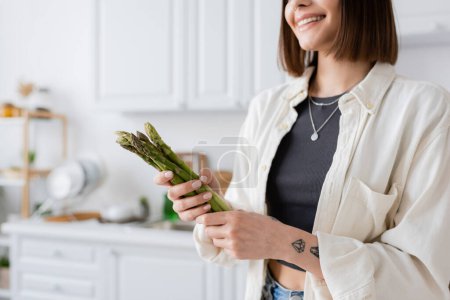 Ausgeschnittene Ansicht tätowierter fröhlicher Frau mit Spargel in der Küche 