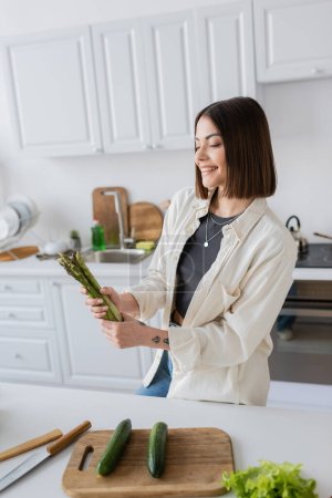 Cheerful young woman holding asparagus near vegetables and cutting board in kitchen 