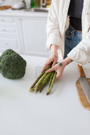 Cropped view of tattooed woman holding asparagus near broccoli and cutting board in kitchen 
