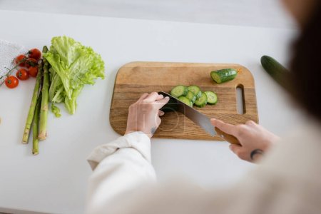 Tätowierte Frau schneidet Gurken in der Nähe von Gemüse in der Küche 