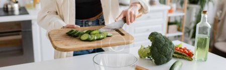 Cropped view of woman holding cut cucumber on cutting board near bowl in kitchen, banner 