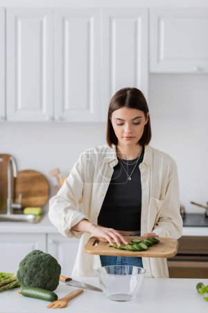 Brunette woman holding sliced cucumber while cooking salad in kitchen 