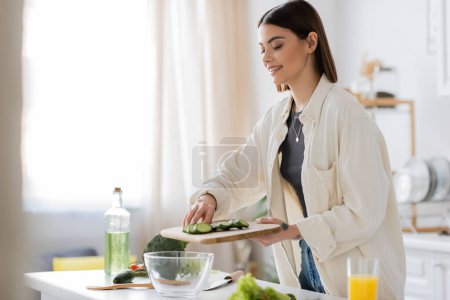 Carefree woman holding cucumber near bowl and vegetables in kitchen 