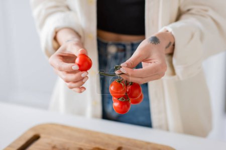Cropped view of blurred woman holding ripe cherry tomatoes near cutting board in kitchen 
