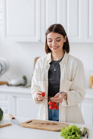 Positive woman holding cherry tomatoes near vegetables and cutting board in kitchen 