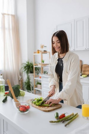 Positive brunette woman cutting lettuce while cooking salad in kitchen 