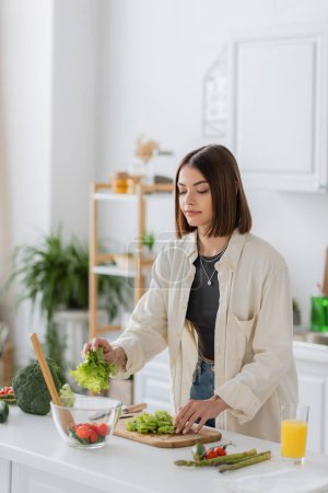 Young brunette woman putting lettuce in bowl while cooking salad in kitchen 