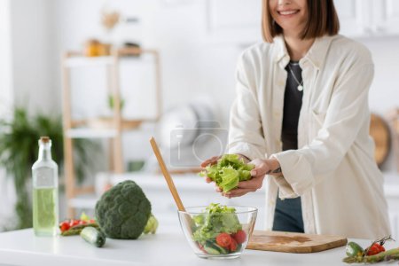 Cropped view of smiling woman putting lettuce in bowl with salad in kitchen 
