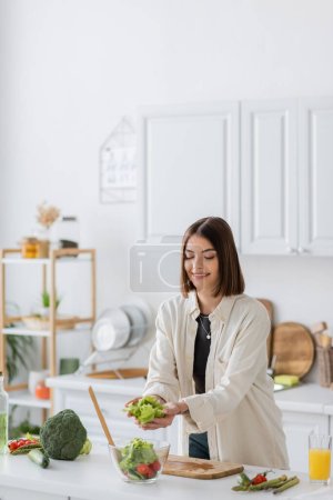 Overjoyed brunette woman putting lettuce in bowl with salad in kitchen 