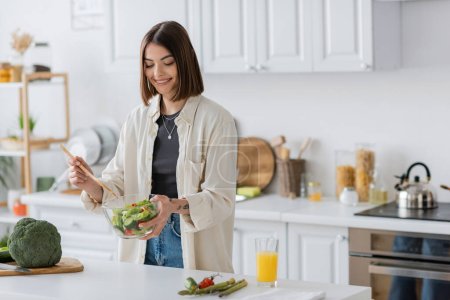 Mujer sonriente sosteniendo tazón con ensalada fresca en la cocina en casa 